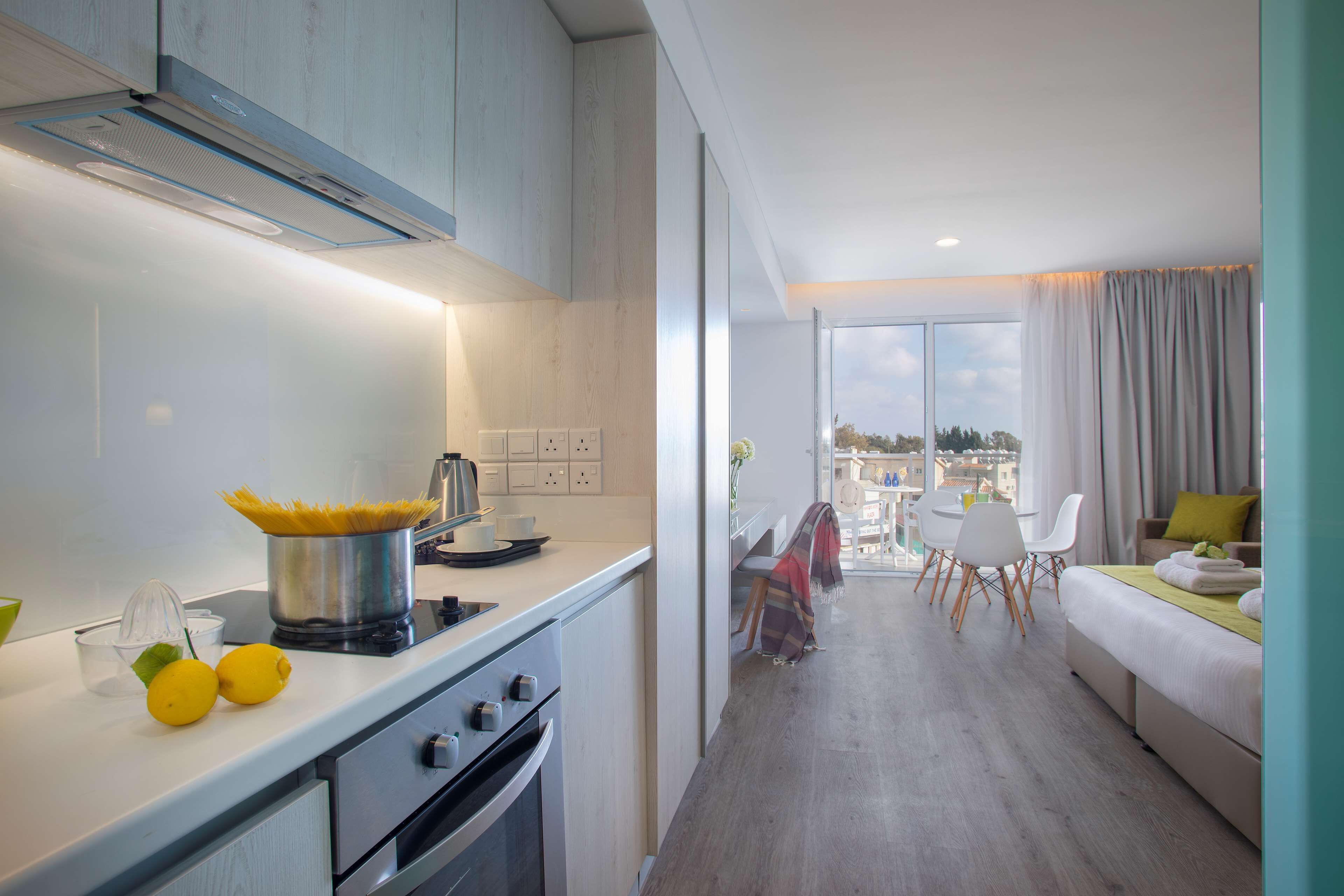 A modern kitchen with a stove, sink, and a pot of spaghetti. There is a dining area with a table and chairs, and a balcony with a view of the city.