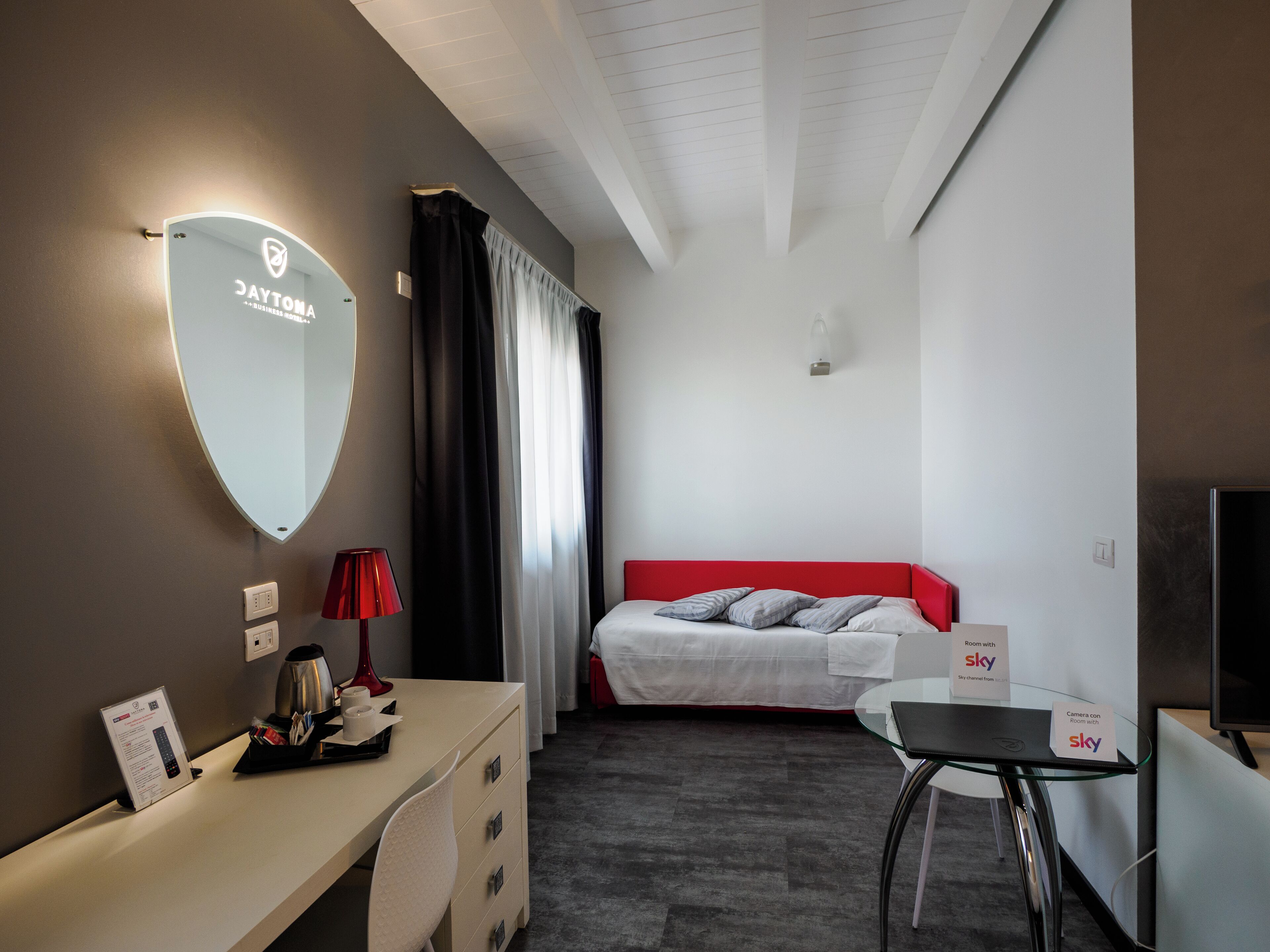 A hotel room with a desk, a bed with a red headboard, a large mirror, and a TV.