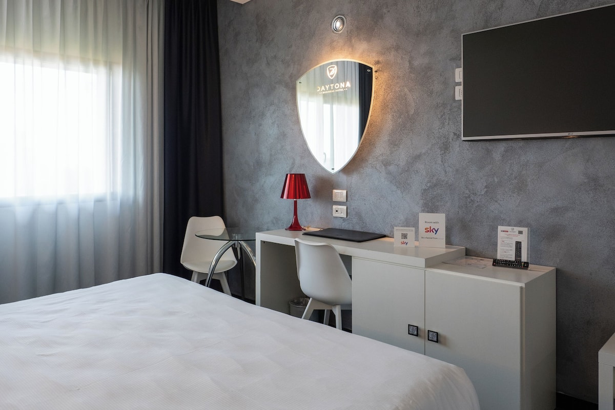 A hotel room with a large bed, a desk with a computer, a red lamp, a mirror, a TV, and a sign indicating Sky room availability.