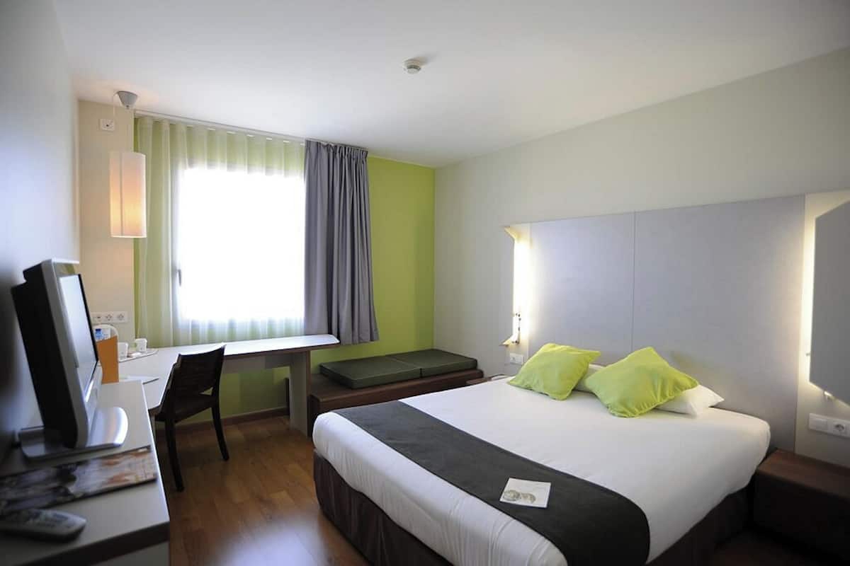 Una habitación de hotel con cama, escritorio, silla y ventana con cortinas.