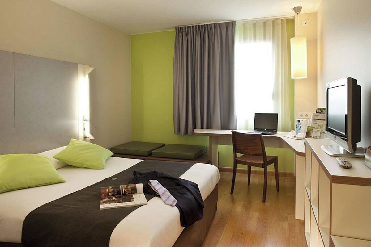 Una habitación de hotel con cama, escritorio, silla, televisor y ventana con cortinas.