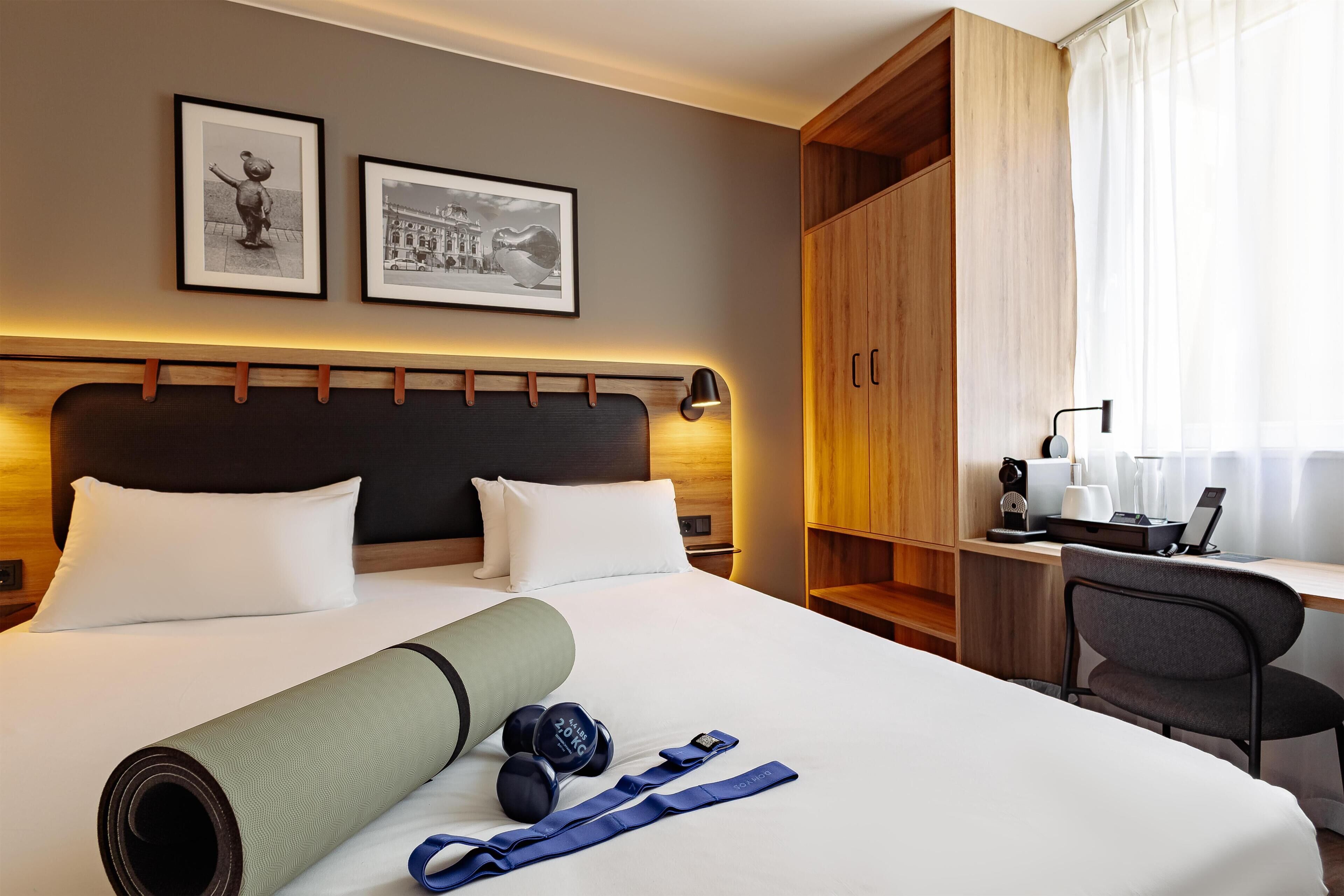 A hotel room with a bed, a desk, a chair, a wardrobe, and two framed pictures on the wall.