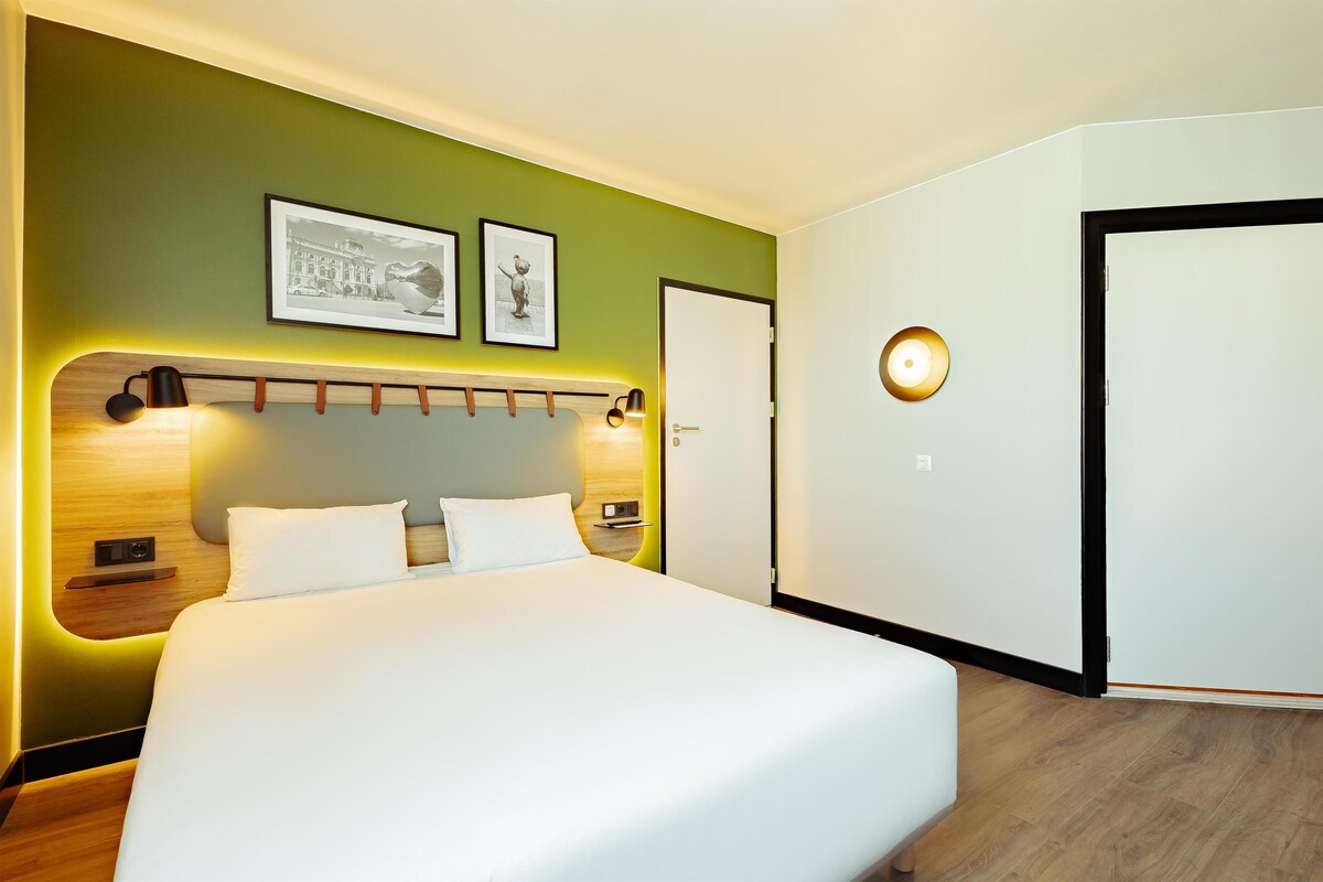 A hotel room with a bed, bedside tables, a wall-mounted lamp, and framed pictures.