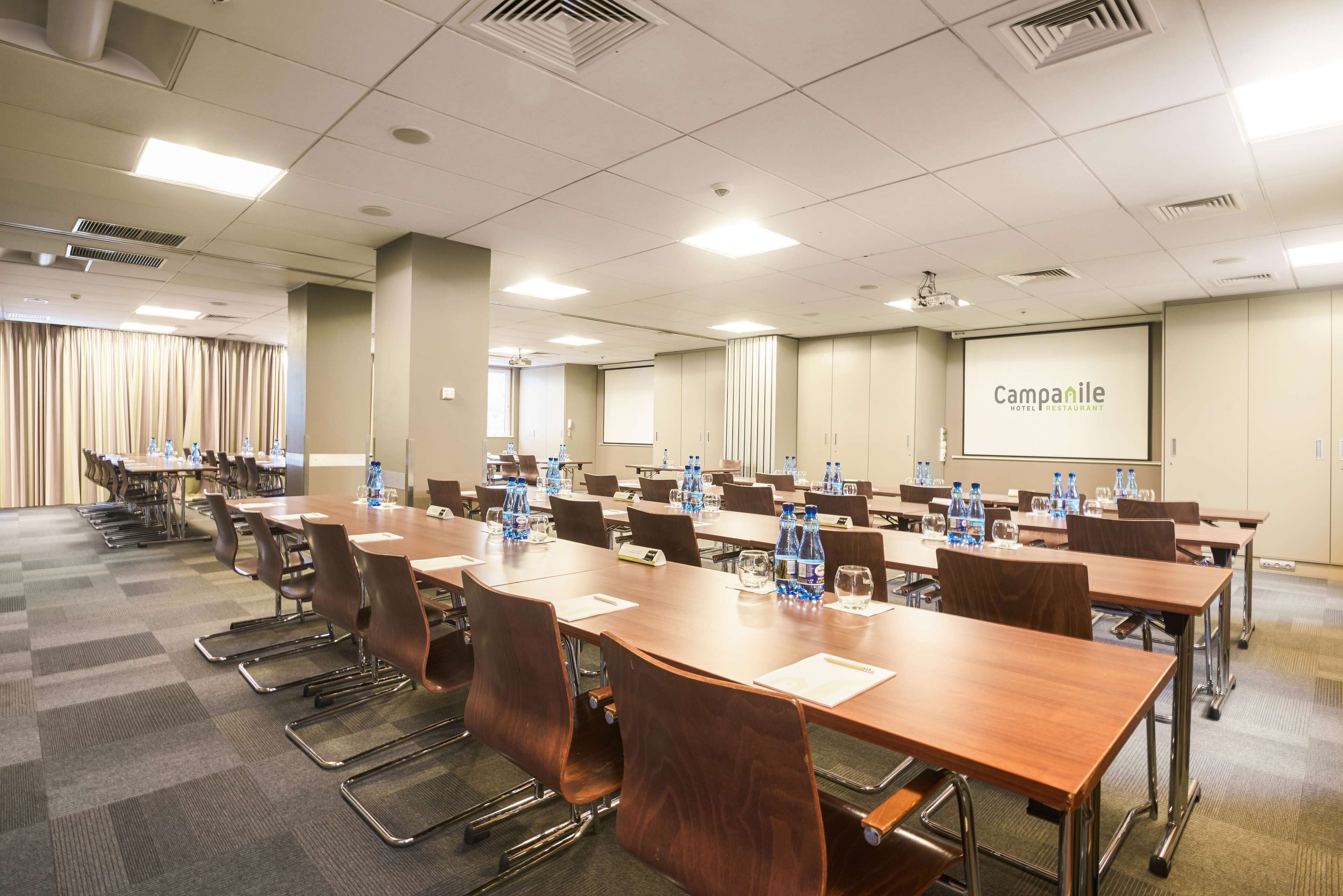 A conference room with multiple tables and chairs, water bottles, and a projector screen displaying "Campanile Hotel Restaurant".