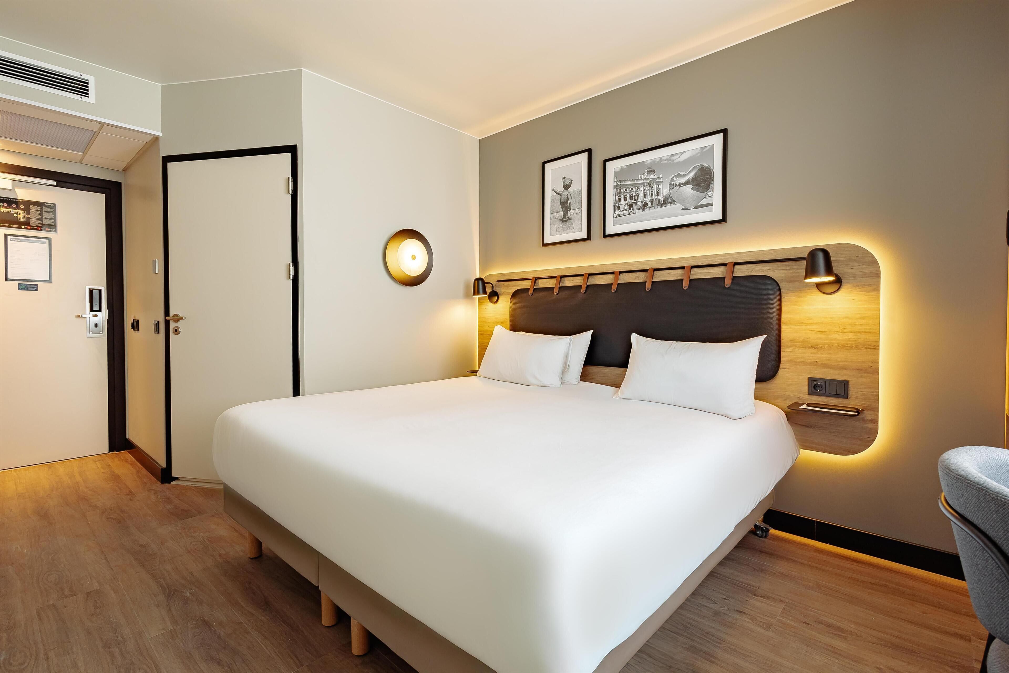 A modern hotel room with a large bed, bedside lamps, a door, and a wall-mounted picture.