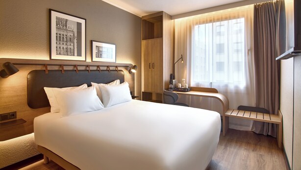 Habitación de hotel con una cama grande, un escritorio con silla, un armario y dos cuadros en la pared.