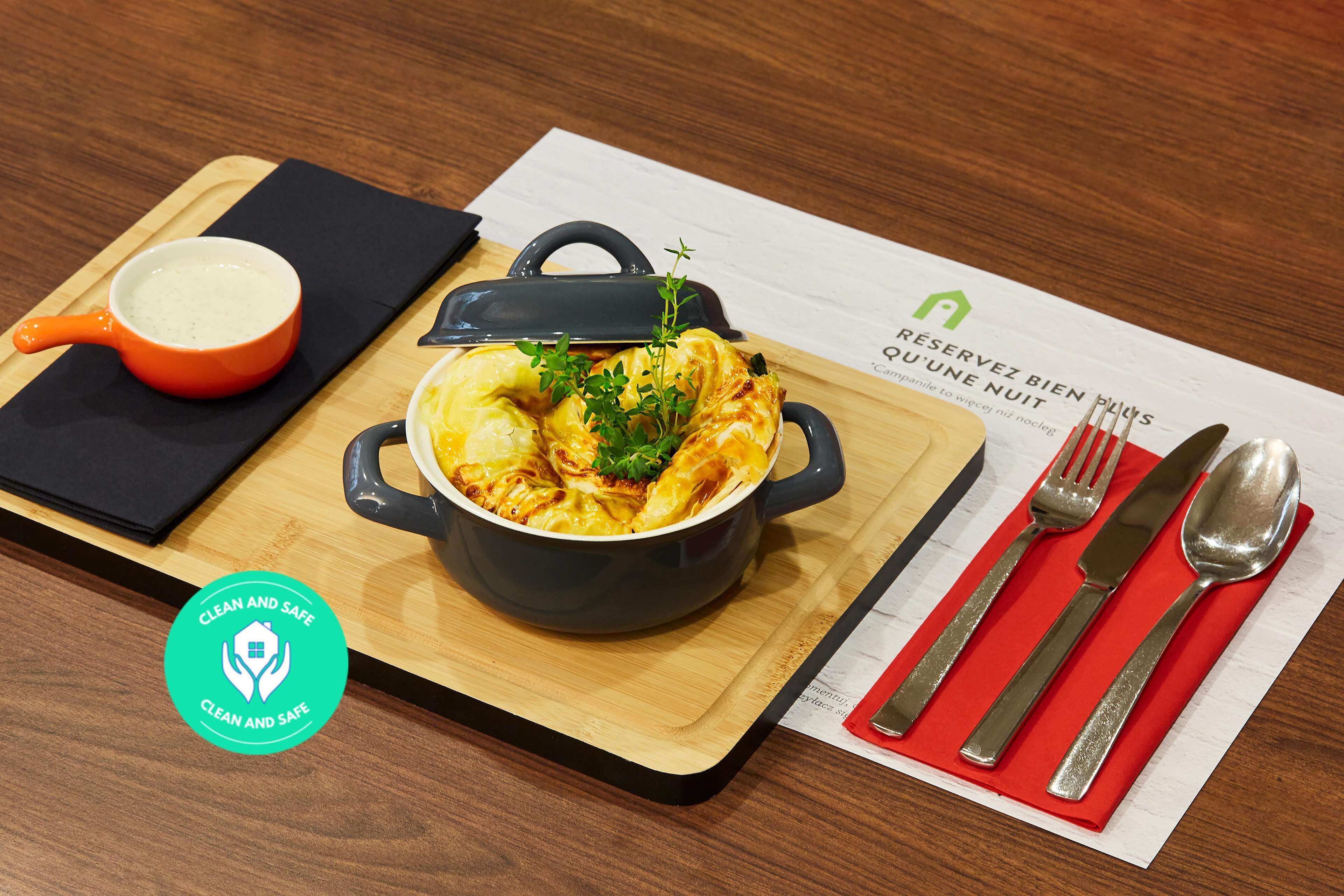 A meal setup with a pot of food, a small orange bowl, and a spoon on a wooden board.