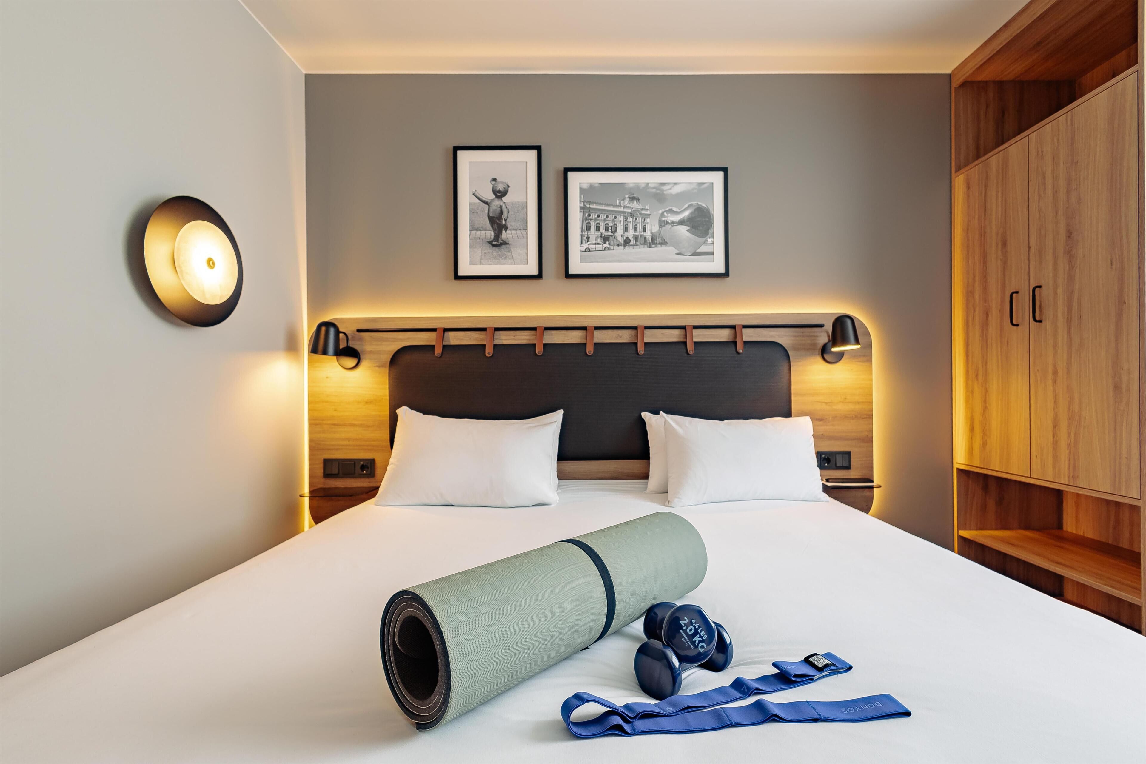 Cama bem arrumada com tapete de yoga, halteres e faixas de resistência em quarto de hotel, com cabeceira, luminárias fixadas na parede e quadros.