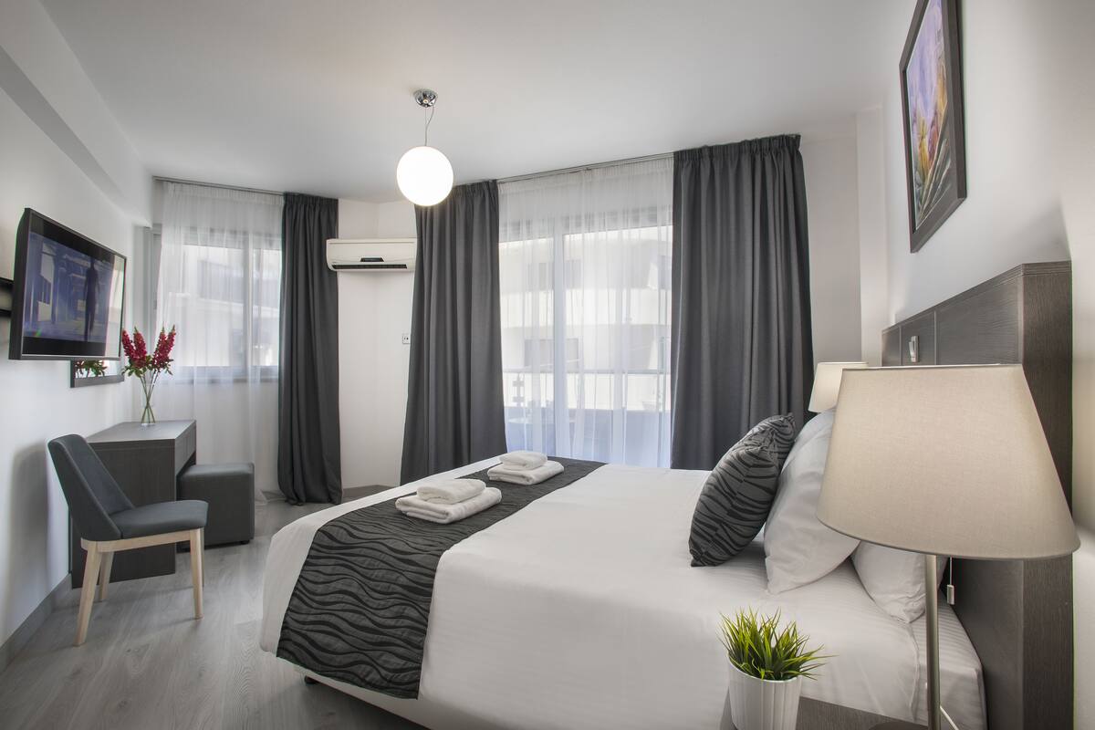 A modern hotel room with a large bed, a desk, a chair, a TV, and a plant.