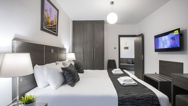 A modern hotel room with a large bed, a bedside table with a lamp, a TV mounted on the wall, and a wardrobe. - Blazer Residence (Larnaca)