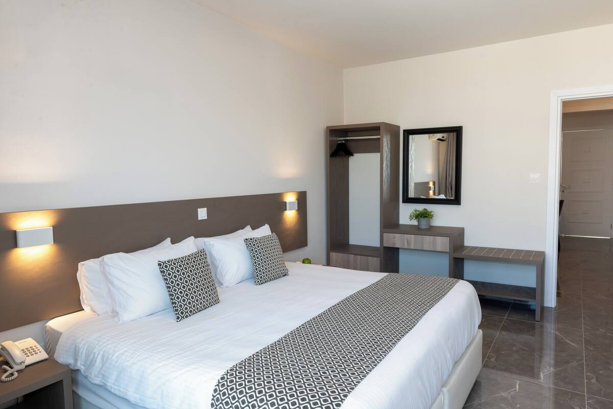 A modern hotel room with a large bed, bedside lamp, a small table with a phone, a mirror, and a bench.