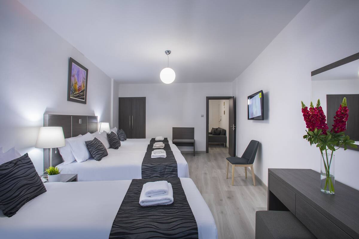 A modern hotel room with two beds, a desk, a chair, a bedside table with a lamp, a vase with flowers, and a painting on the wall.