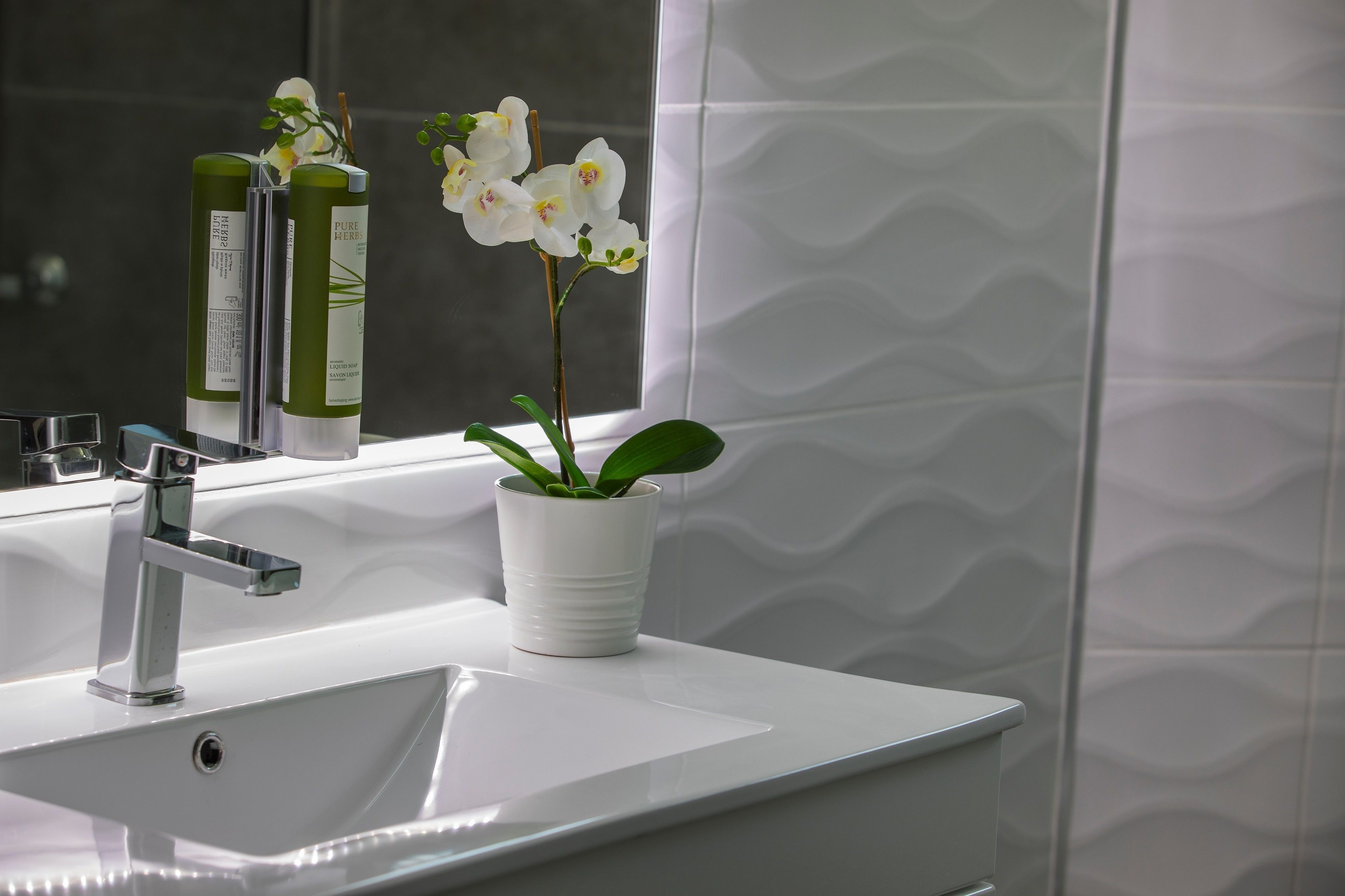 Une salle de bain avec un lavabo blanc, un miroir et une fenêtre ornée d’orchidées blanches.