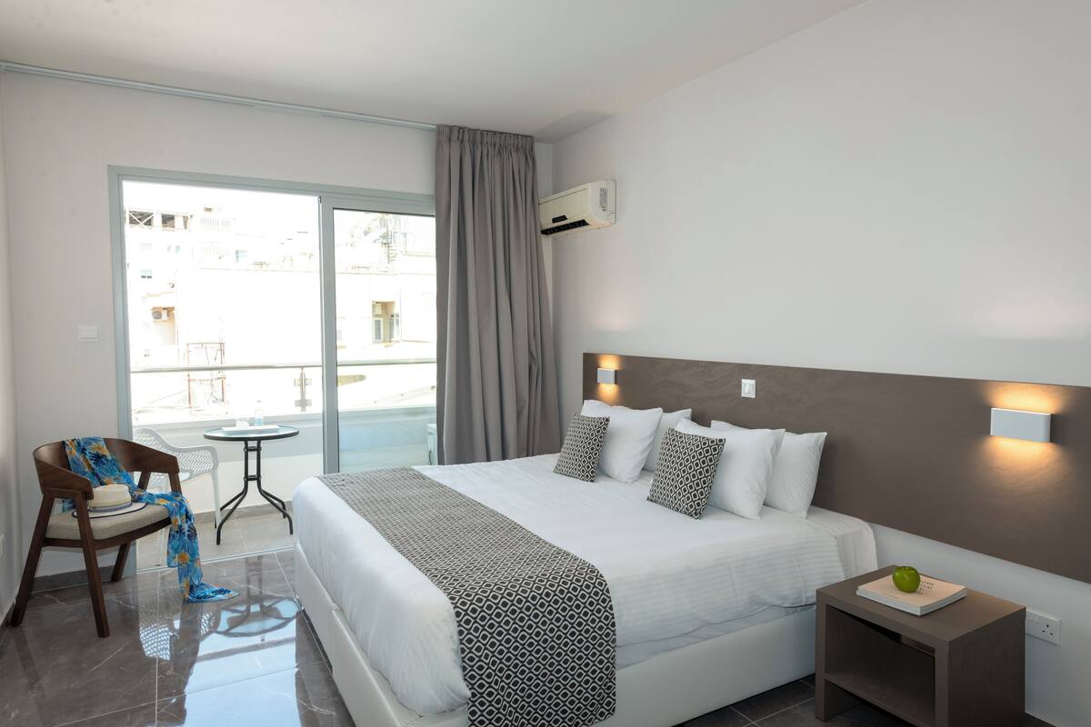 A hotel room with a bed, a chair, a small table, and a view of the outside.