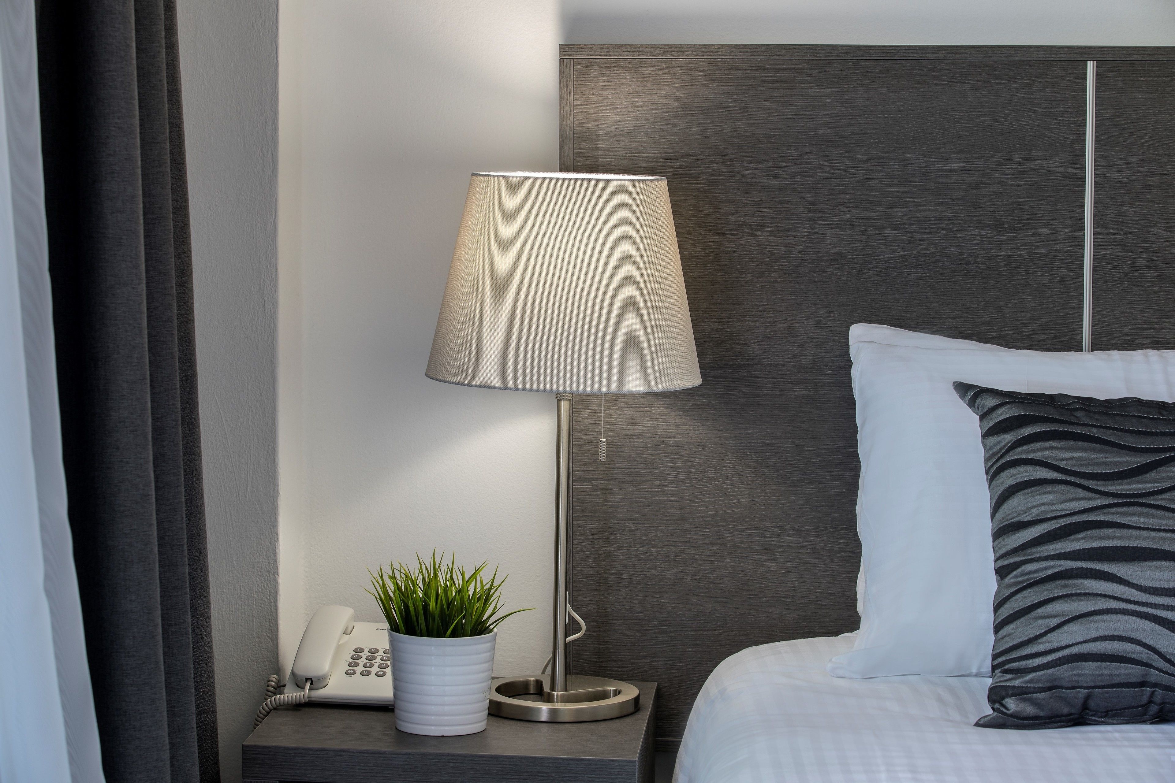 A neatly made bed with a white and grey striped pillow, a grey headboard, a bedside table with a lamp, a phone, and a potted plant.