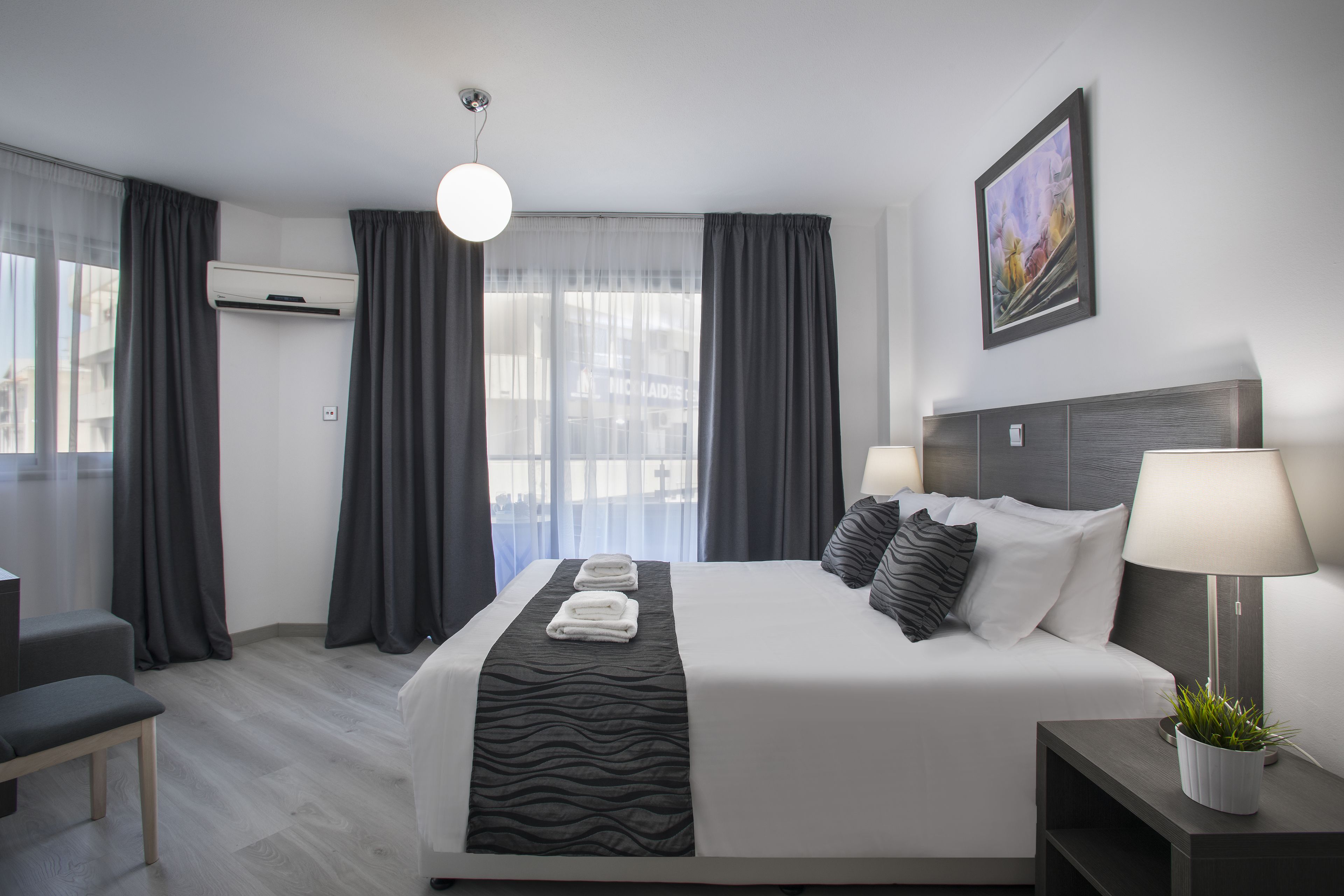 A modern hotel room with a large bed, two bedside lamps, a potted plant, and a city view.