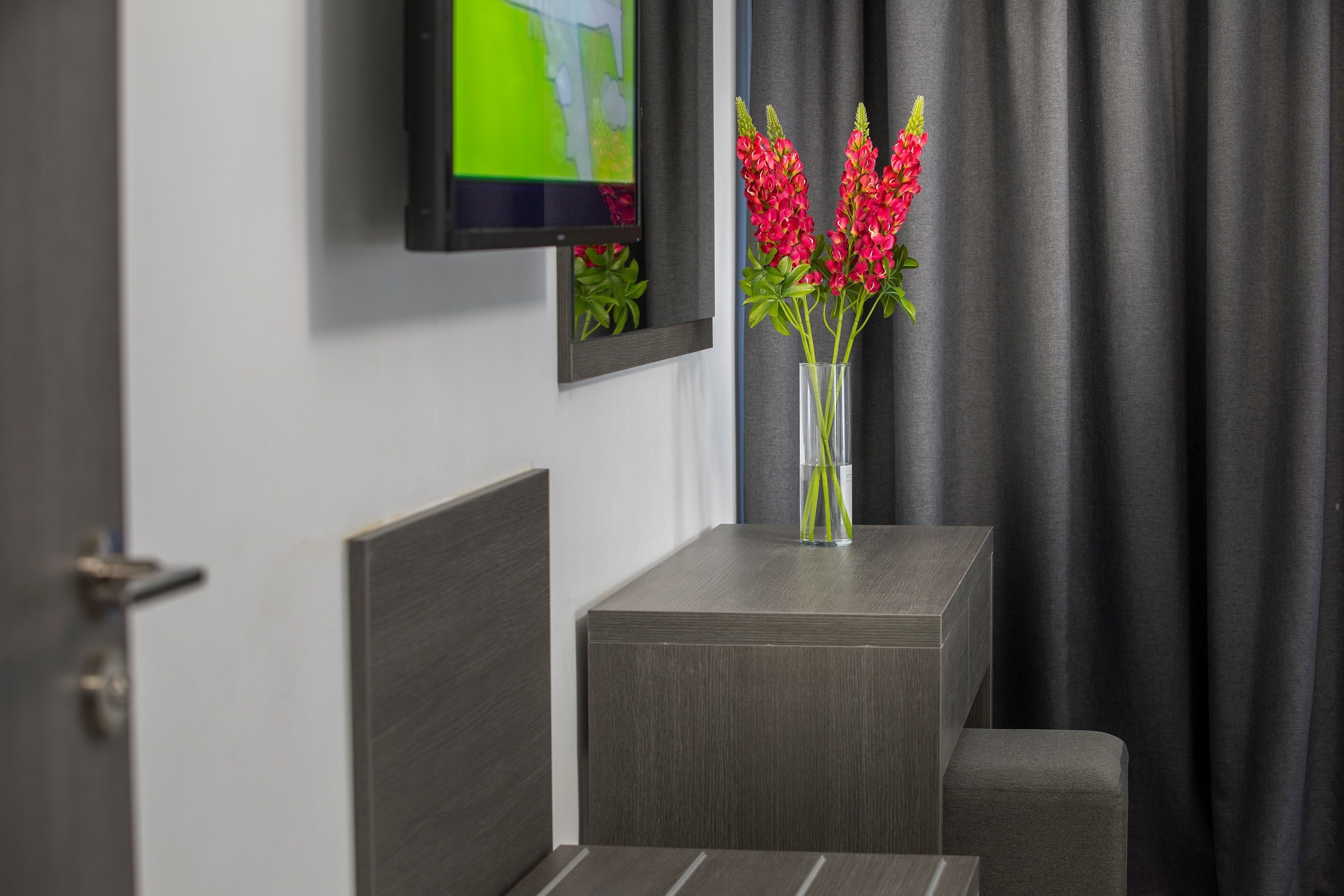 A modern hotel room with a flat-screen TV mounted on the wall, a small table with a vase of red flowers, and a grey curtain.