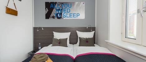Un lit double recouvert d’une couverture matelassée de couleur sombre, un magazine et une casquette marron posés dessus. Un tableau est accroché au mur, sur lequel on peut lire : « TOUT CE DONT VOUS AVEZ BESOIN POUR DORMIR ».