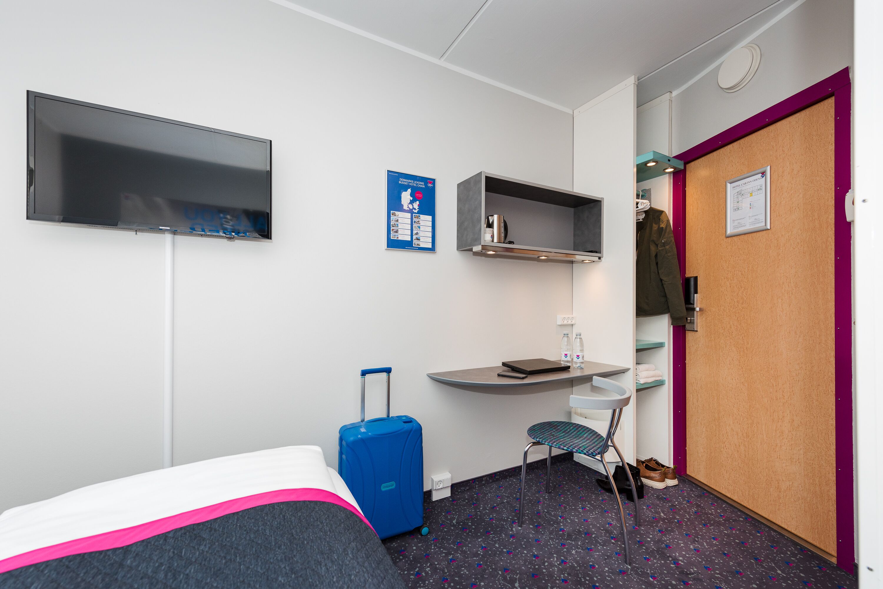 A hotel room with a bed, a desk, a chair, a TV, a coat rack, and a door.