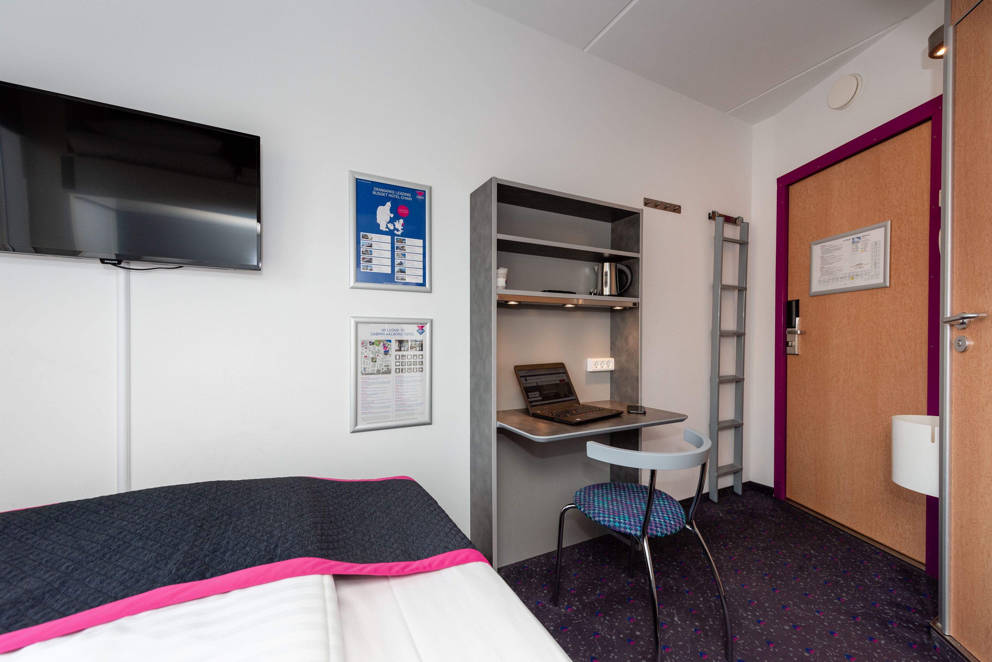 A hotel room with a bed, a desk with a laptop, a chair, a TV mounted on the wall, and a door with a sign.