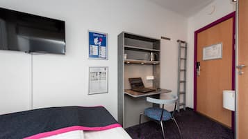 A hotel room with a bed, a desk with a laptop, a chair, a TV mounted on the wall, and a door with a sign.