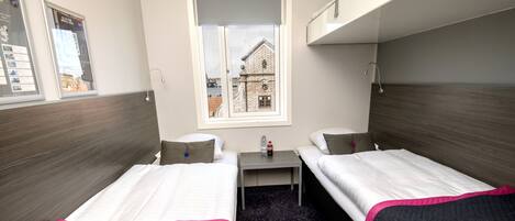 Una habitación de hotel pequeña con dos camas individuales, una mesita, una ventana con vistas a un edificio y una cama litera encima.
