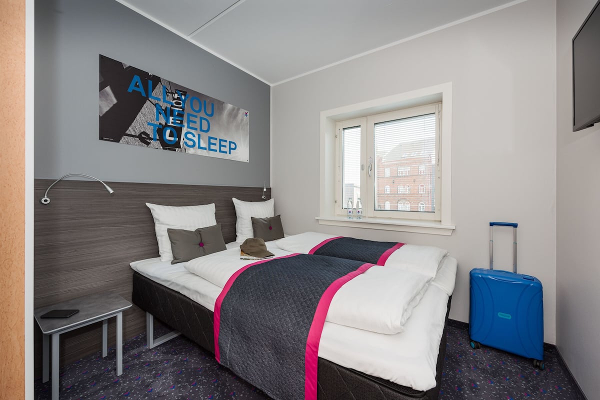 A hotel room with two beds, a window with blinds, a blue suitcase, and a poster on the wall.