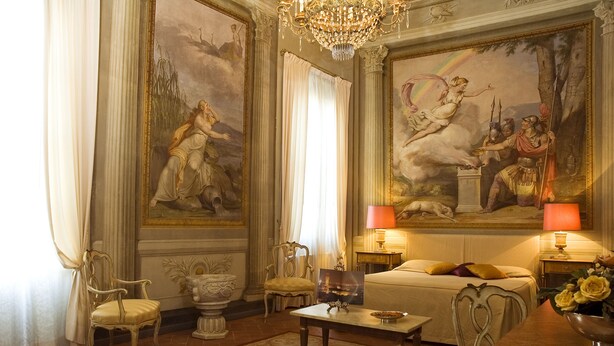 Une chambre luxueuse avec un grand lit, des fauteuils richement décorés, un lustre et des tableaux classiques aux murs.