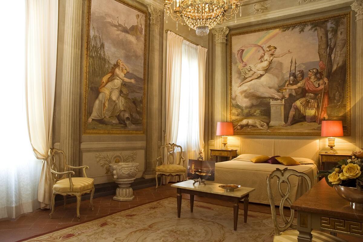 Una habitación lujosa con una cama grande, sillas ornamentadas, una araña de cristal y pinturas clásicas en las paredes.