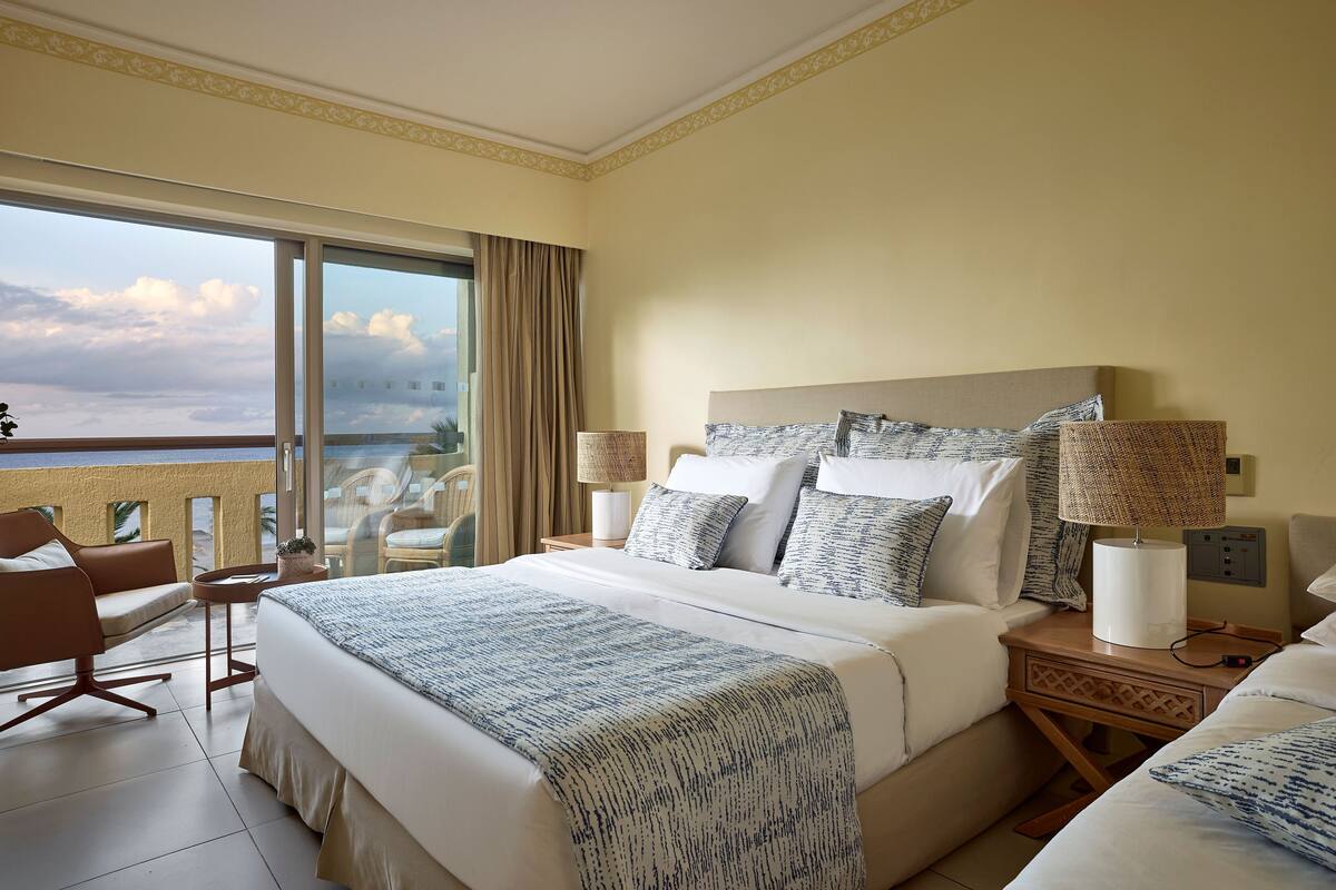 Ein Hotelzimmer mit einem großen Bett, zwei Nachttischen, einem Sessel und Meerblick.