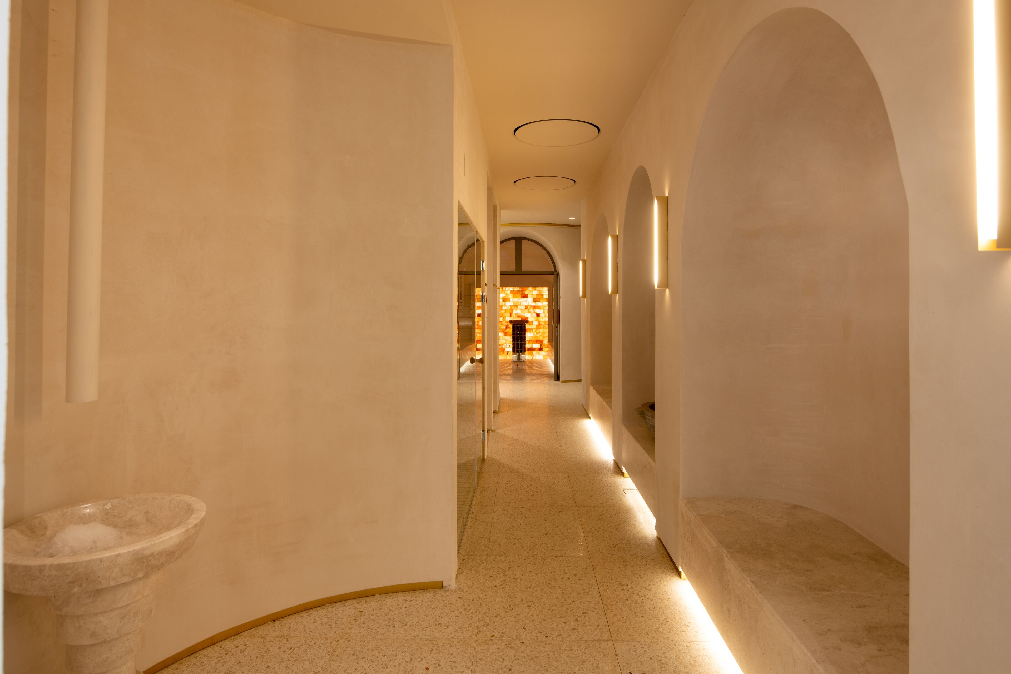 A corridor with arched ceilings, a stone sink, and a warm light source at the end.