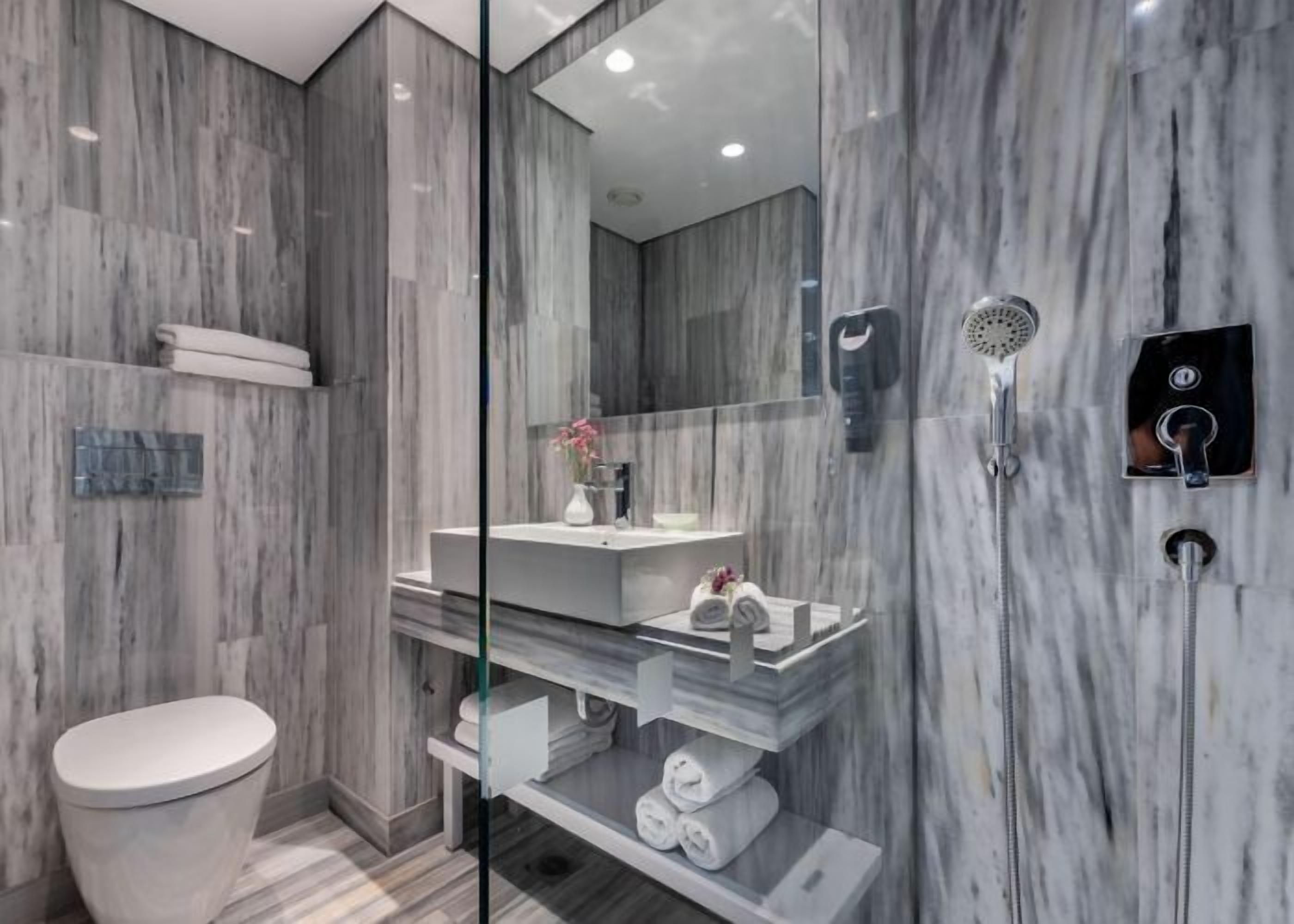 A modern bathroom with a marble wall, a glass shower enclosure, a toilet, and a vanity with a mirror.