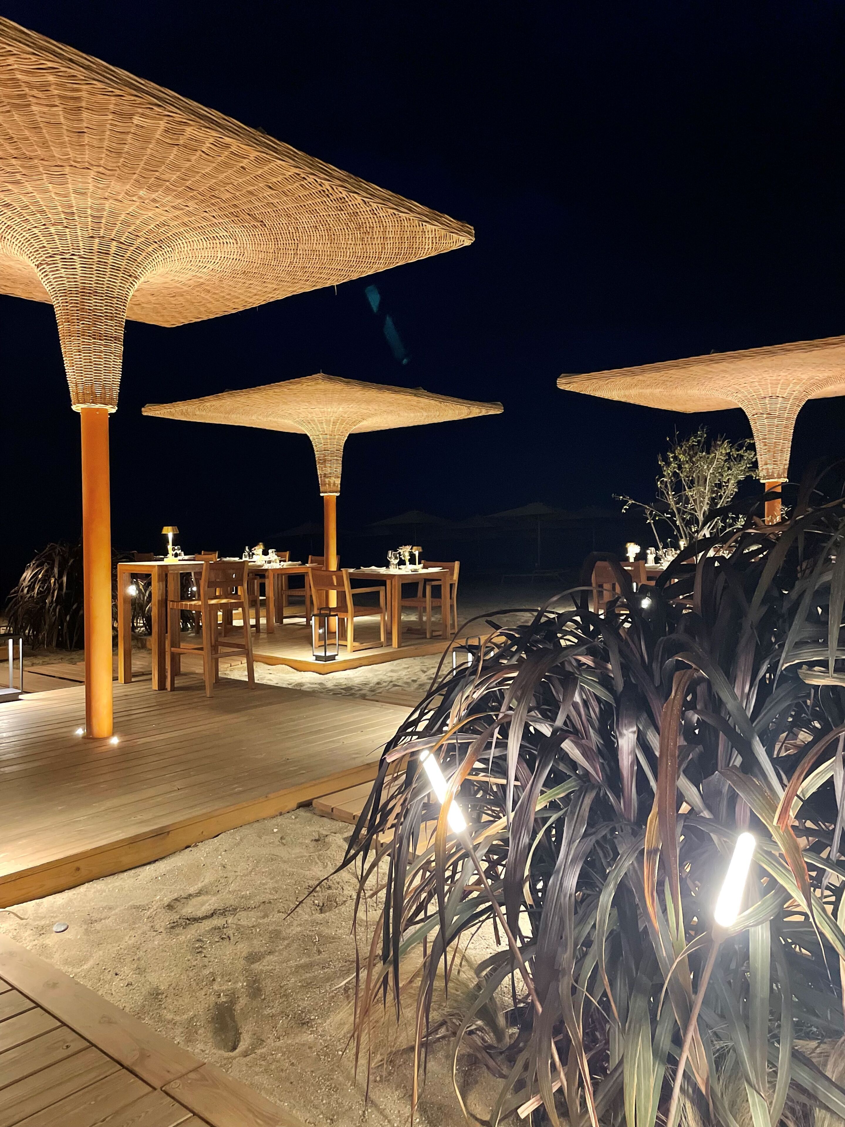 An outdoor dining area with wooden tables and chairs under large woven umbrellas, set on a sandy deck.