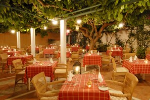 An outdoor dining area with tables covered in red checkered tablecloths, wicker chairs, and candles providing ambient lighting.