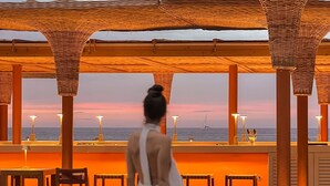Una terraza de madera con vista al mar, donde aparece una mujer con un vestido blanco.