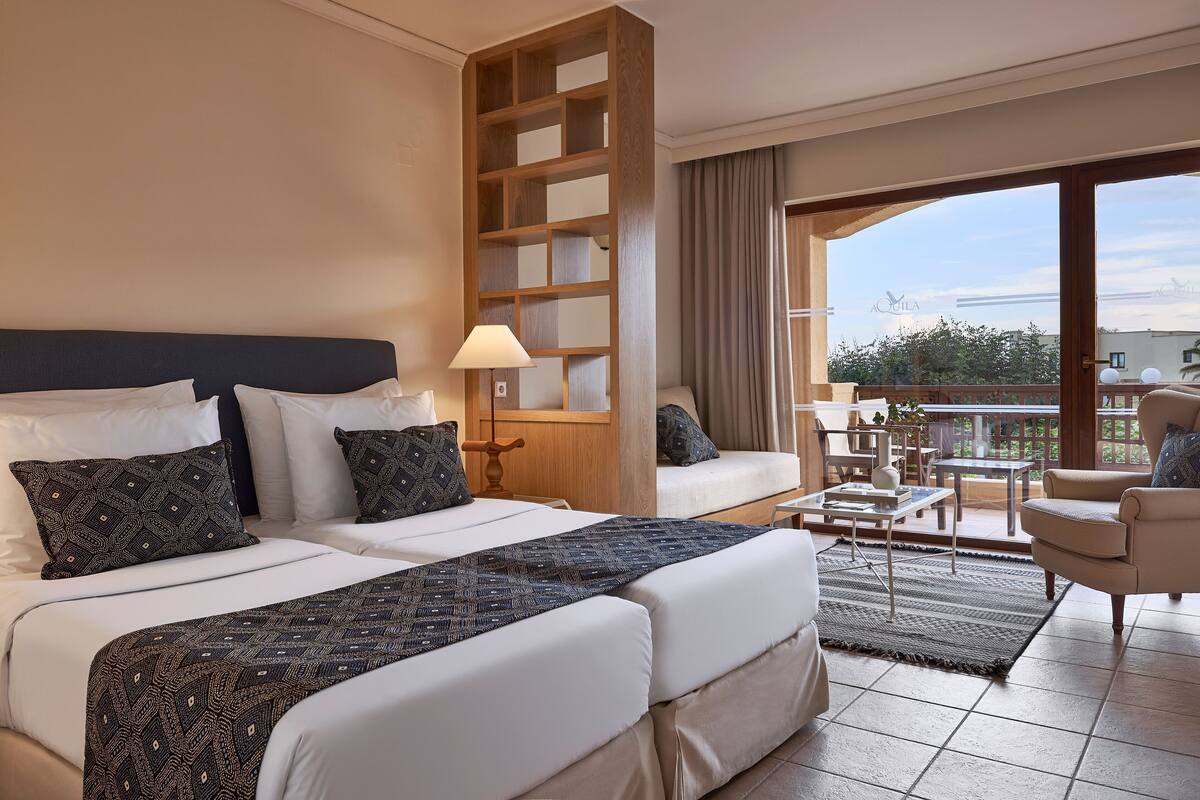 Ein Hotelzimmer mit einem großen Bett, einer Couch, einem Balkon mit Tisch und Stühlen sowie Blick auf die Stadt.