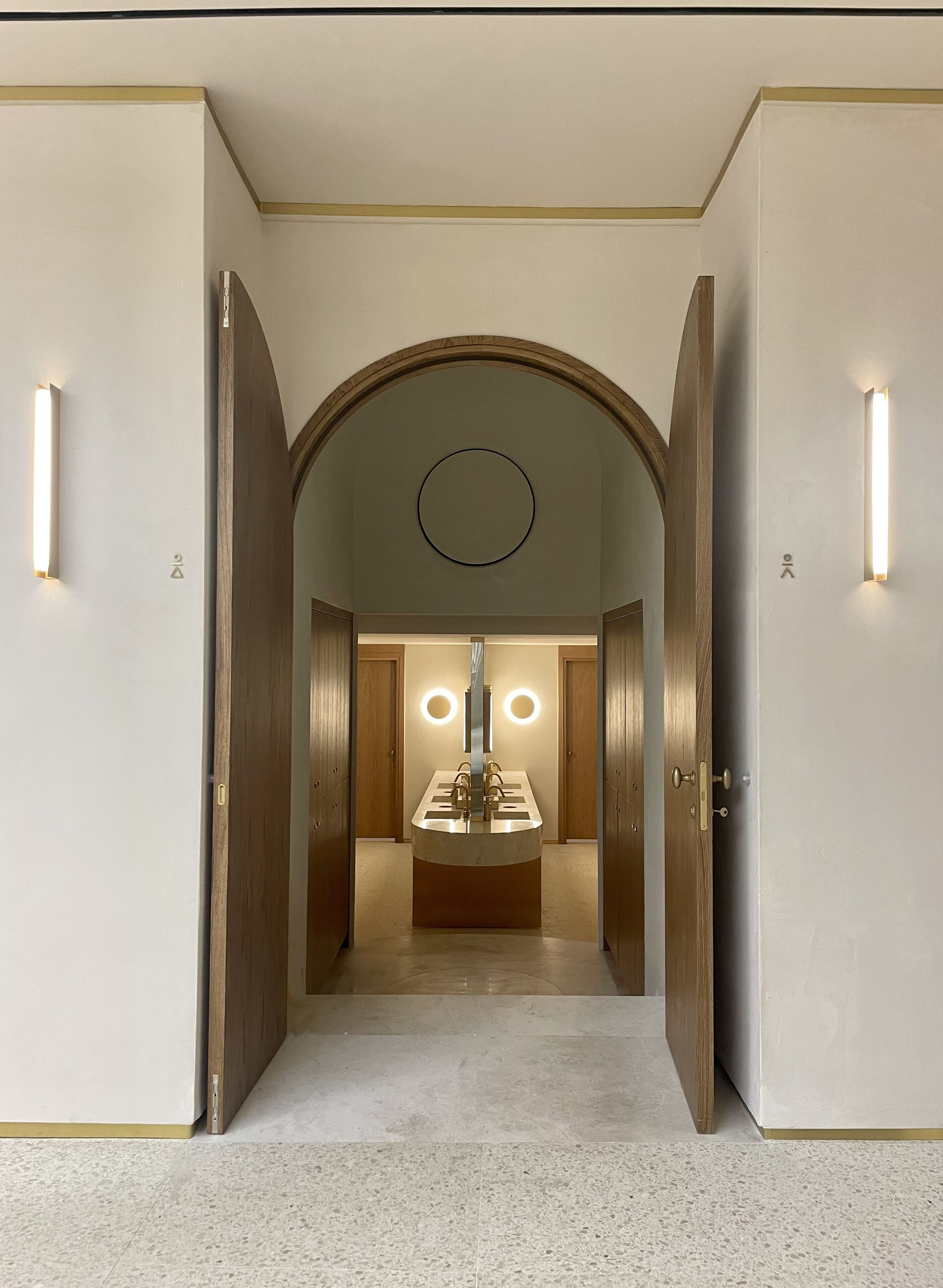 Pasillo de hotel con una zona de tocador central que cuenta con un lavabo y espejos, flanqueado por puertas de madera e iluminado por apliques de pared.