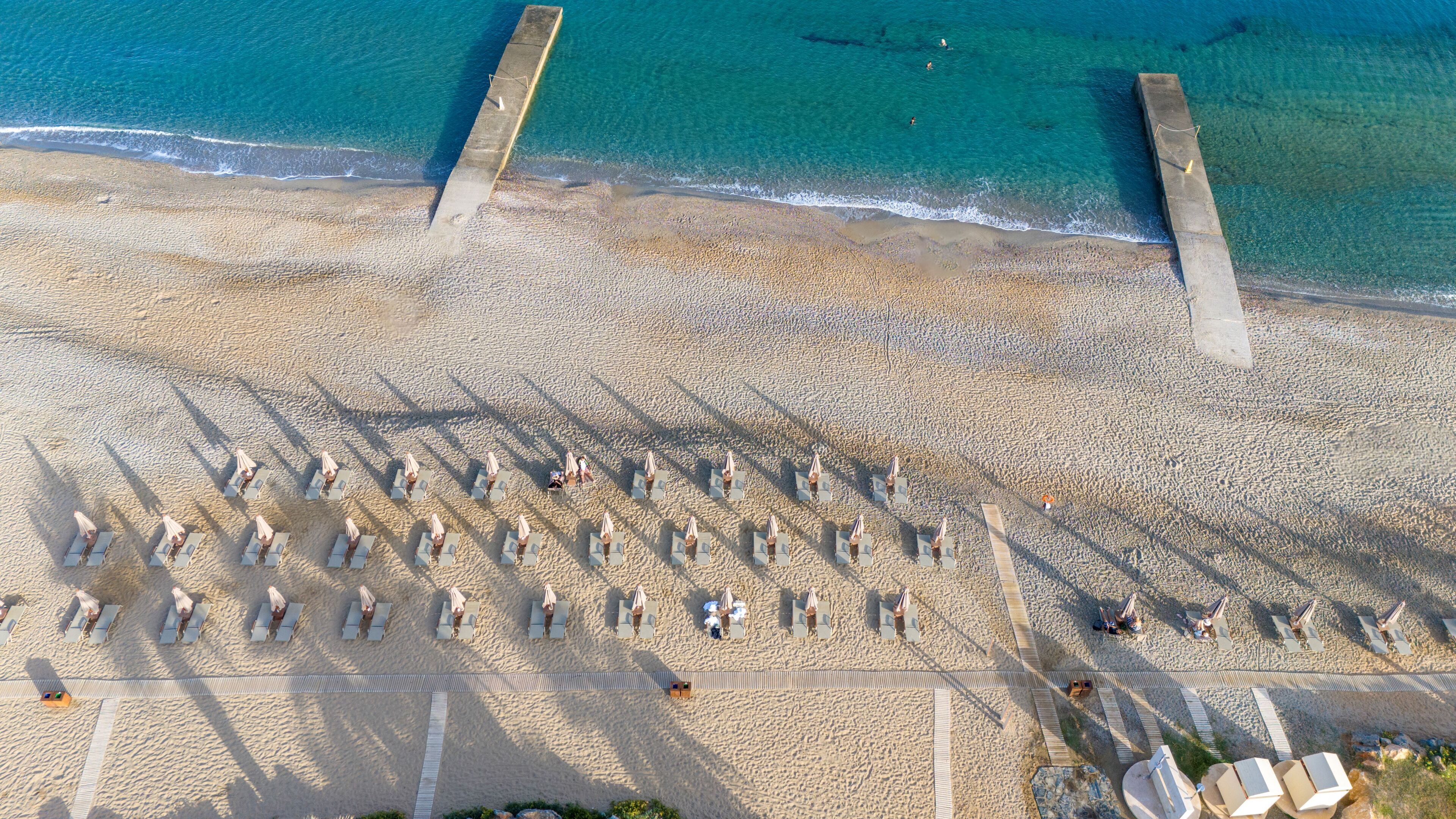 An aerial view of a beach with rows of sun loungers and umbrellas, a concrete pier, and clear blue water.