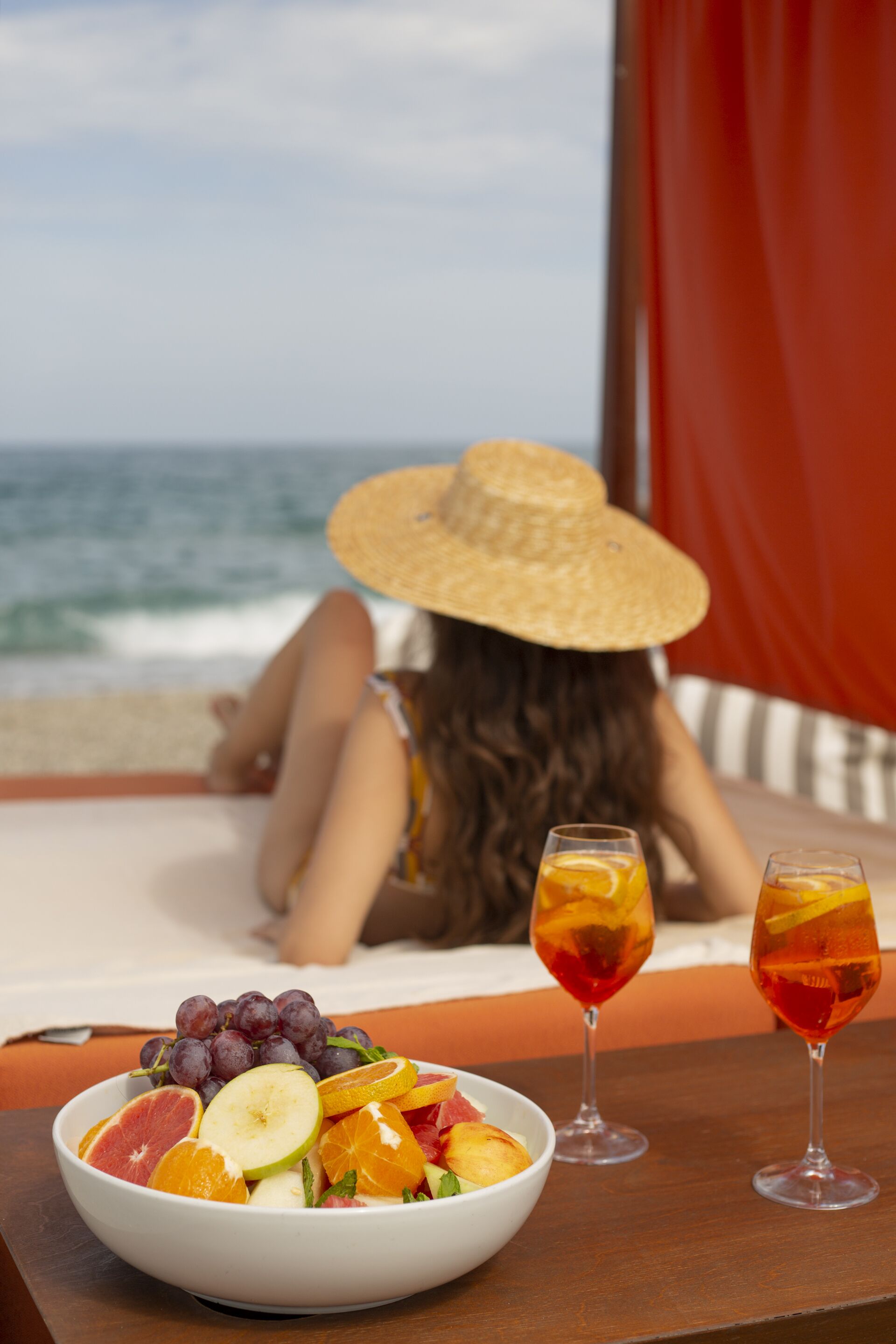 A beach scene with a woman in a straw hat, a bowl of fruit, and two glasses of drink on a wooden table.