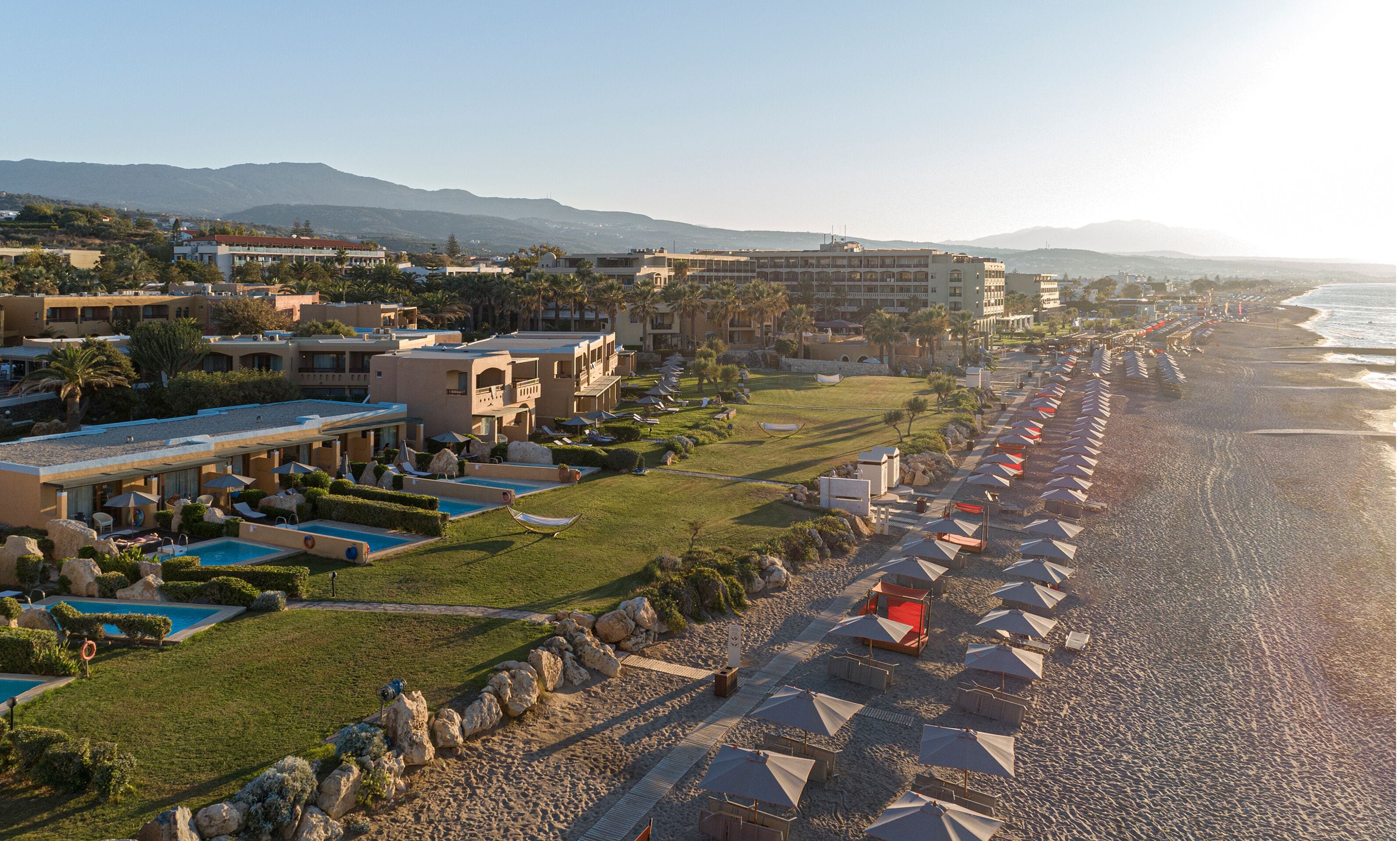 A resort with multiple swimming pools, a beach with umbrellas, and a view of mountains in the background.