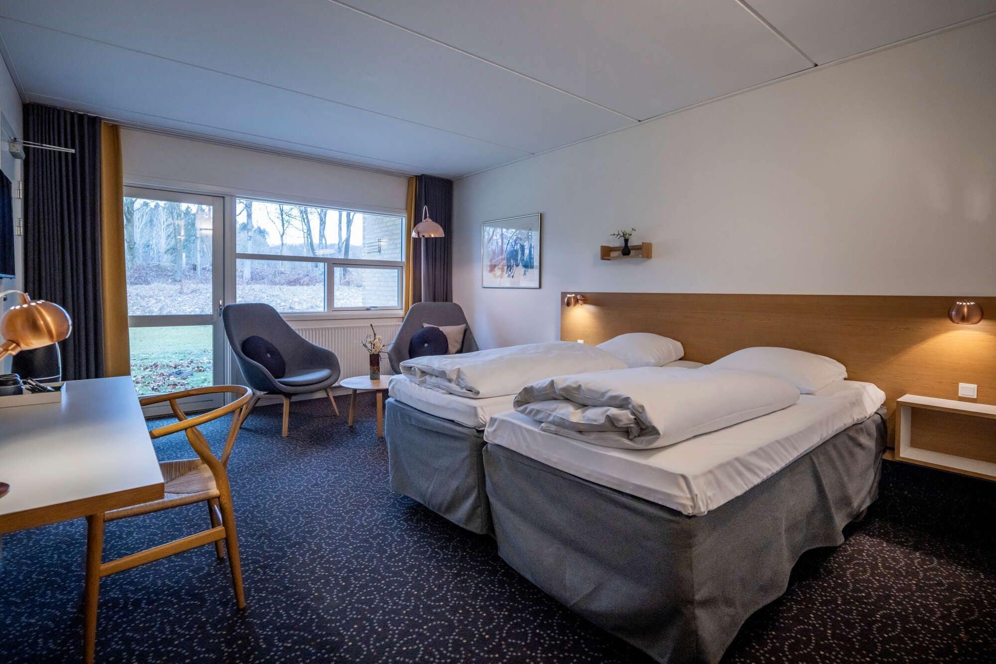 A hotel room with two beds, a desk, a chair, and a large window offering a view of trees.