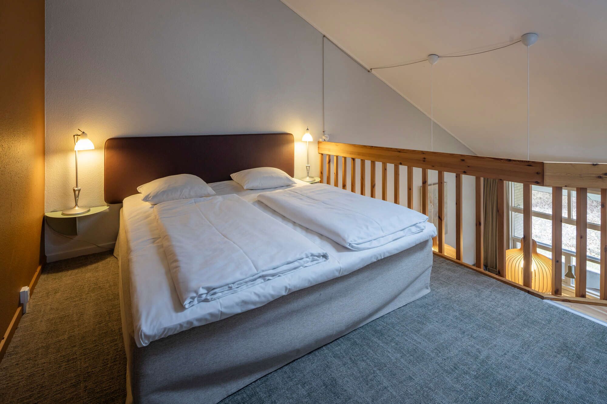 A bedroom with a bed, two bedside tables with lamps, a wooden headboard, and a view of the outdoors through a window.