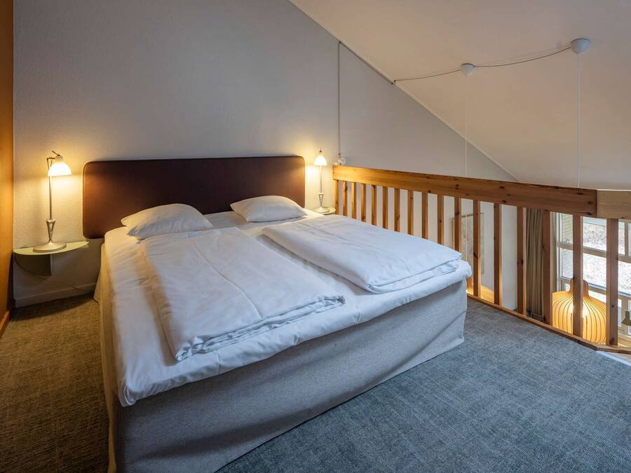 Un dormitorio con cama, dos mesitas de noche con lámparas, cabecera de madera y vistas al exterior a través de una ventana.