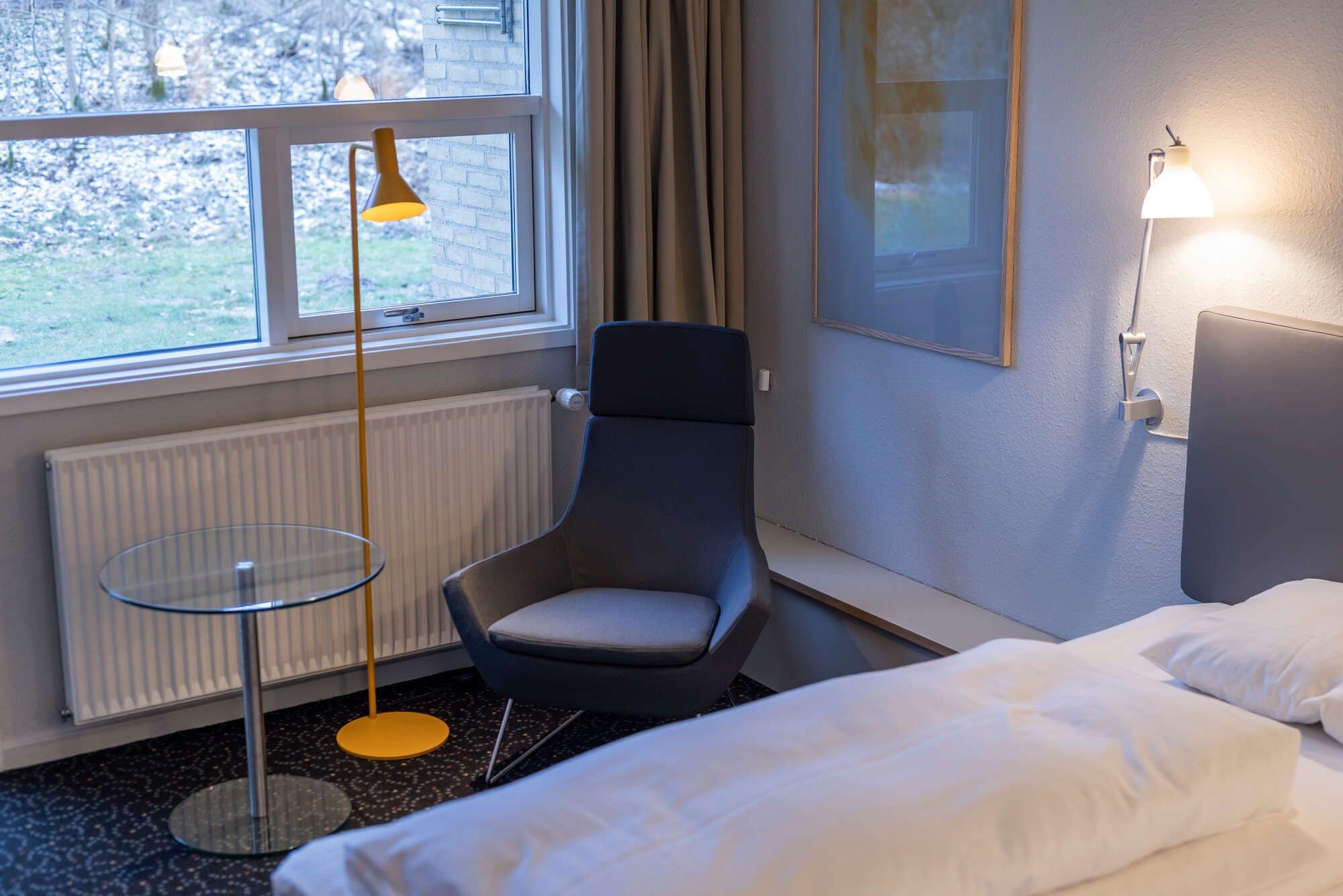 A modern hotel room with a single bed, a chair, a small table, and a window with curtains.