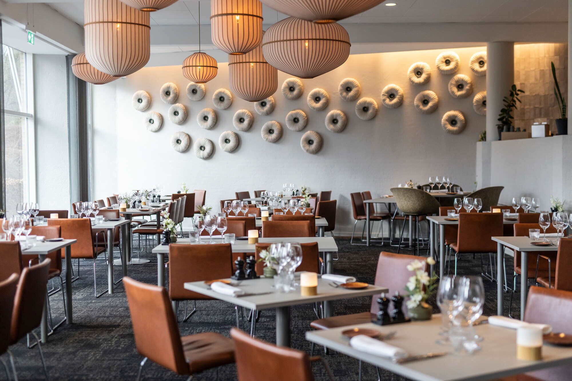 Un interior de restaurante moderno con mesas redondas, sillas de cuero marrón y una pared decorativa con motivos circulares.