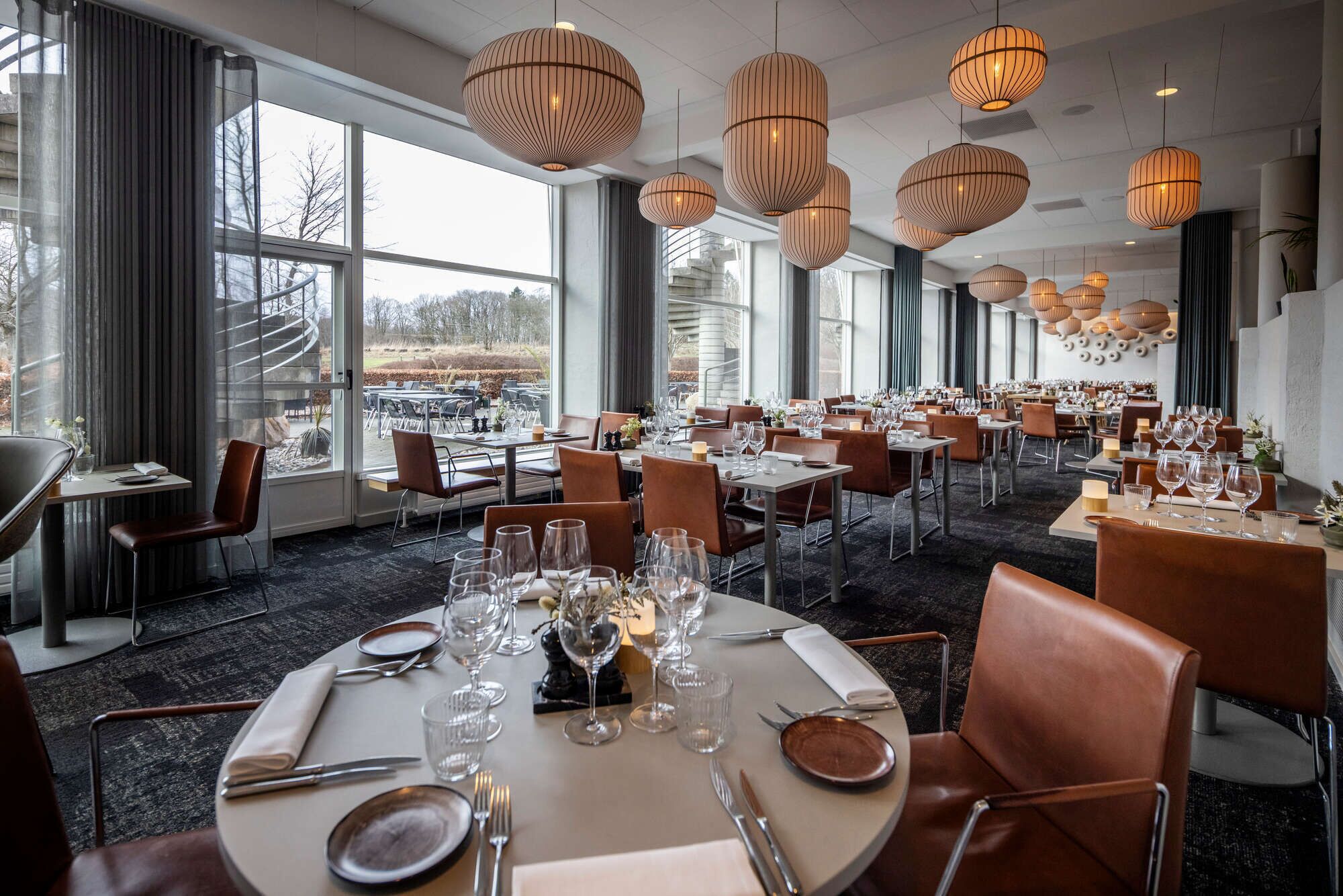A restaurant with round tables set for dining, large windows, and hanging pendant lights.