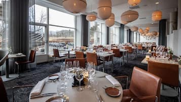 A restaurant with round tables set for dining, large windows, and hanging pendant lights.