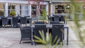 An outdoor seating area with tables and chairs, surrounded by decorative plants and a building with patterned windows. - Comwell Rebild Bakker (Skorping)