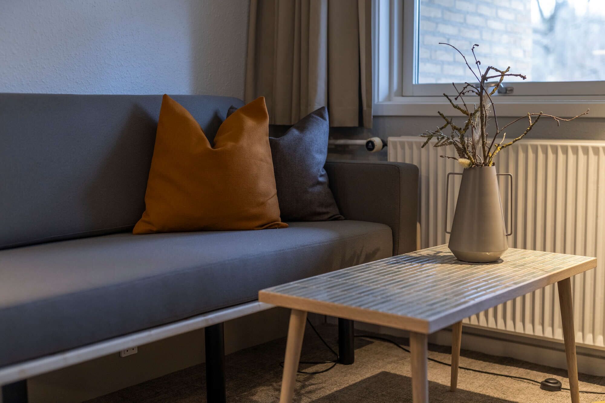 En moderne stue med en grå sofa, et lille træbord og en vase med grene.
