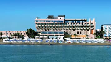 Un hotel de varios pisos, una playa al frente y varias sombrillas y sillas de descanso.